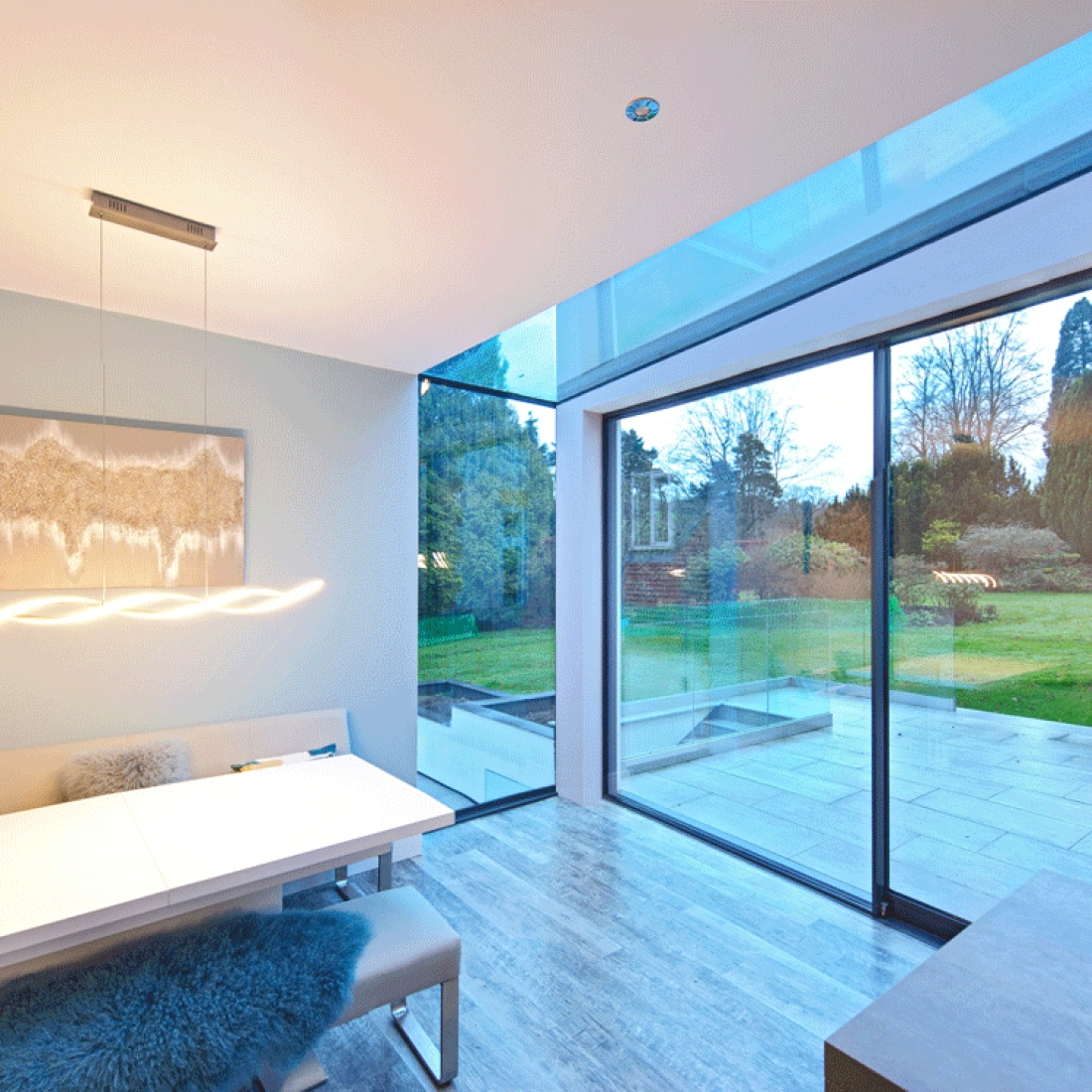 Un intérieur moderne d’une salle à manger avec une grande fenêtre donnant sur un jardin verdoyant. Une table blanche élégante est entourée de sièges confortables. Des lumières suspendues ajoutent une ambiance chaleureuse.