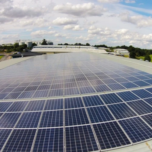 A rooftop covered with numerous solar panels under a partly cloudy sky. In the background, a landscape features green fields and buildings.