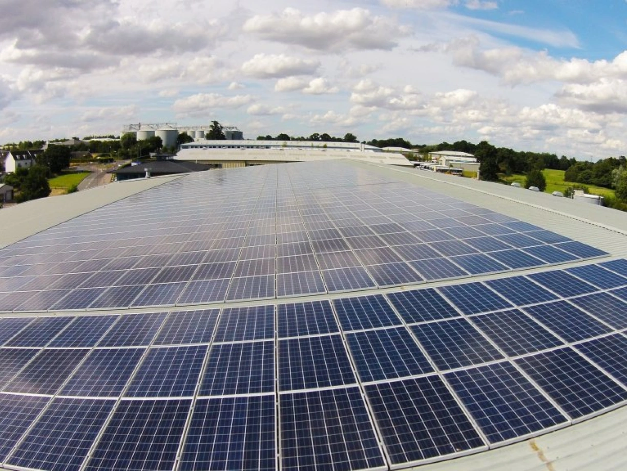 Solaranlage auf einem Dach mit vielen Photovoltaikmodulen. Im Hintergrund sind Gebäude und ein bewölkter Himmel zu sehen.