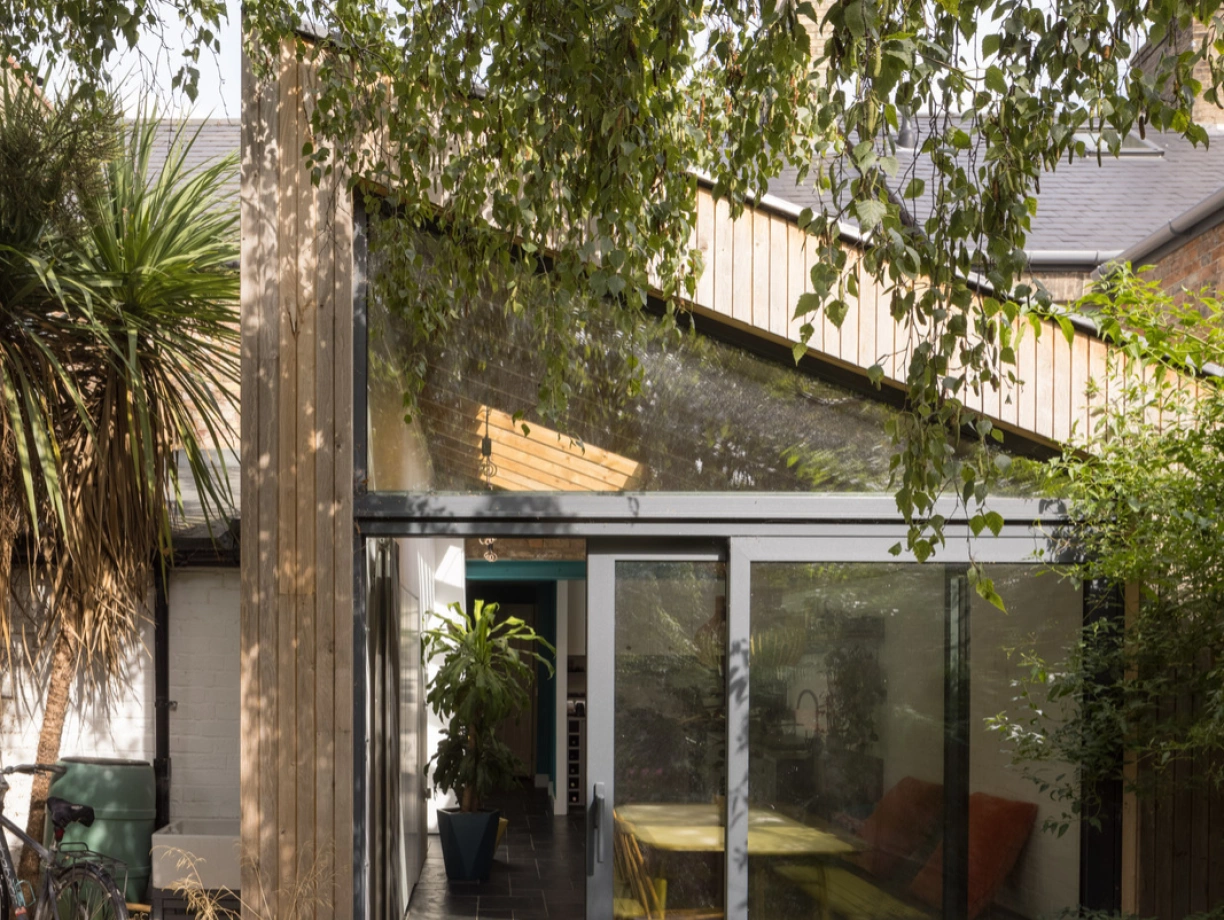 A modern outdoor space with a glass wall connecting to a partly covered patio. Bright yellow tables and chairs contrast with lush greenery and a bicycle parked nearby. Sunlight filters through the leaves, creating a serene atmosphere.