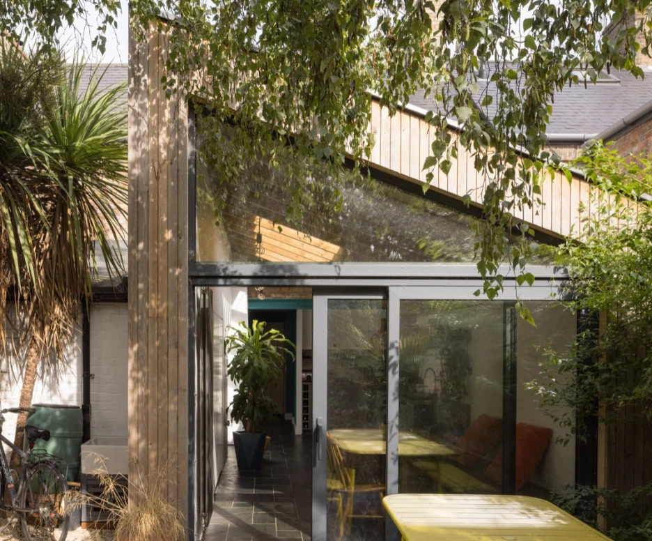 A modern outdoor space with a glass wall connecting to a partly covered patio. Bright yellow tables and chairs contrast with lush greenery and a bicycle parked nearby. Sunlight filters through the leaves, creating a serene atmosphere.