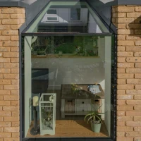 A cozy nook with a glass window surrounded by brick walls. Inside, there is a small table with a plant and decorative items, along with a comfortable chair. The reflection of greenery and buildings can be seen in the glass.