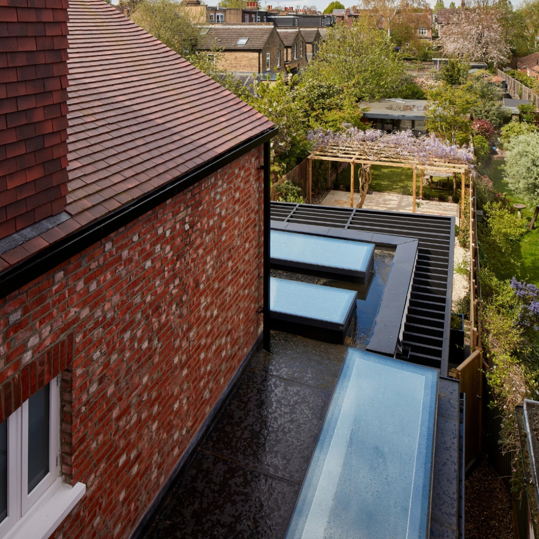 Aerial view of a modern backyard featuring a sleek black decking area with two rectangular pools. Surrounded by greenery, trees, and a pergola adorned with climbing plants. A red-bricked house is visible in the foreground with a sloped roof.