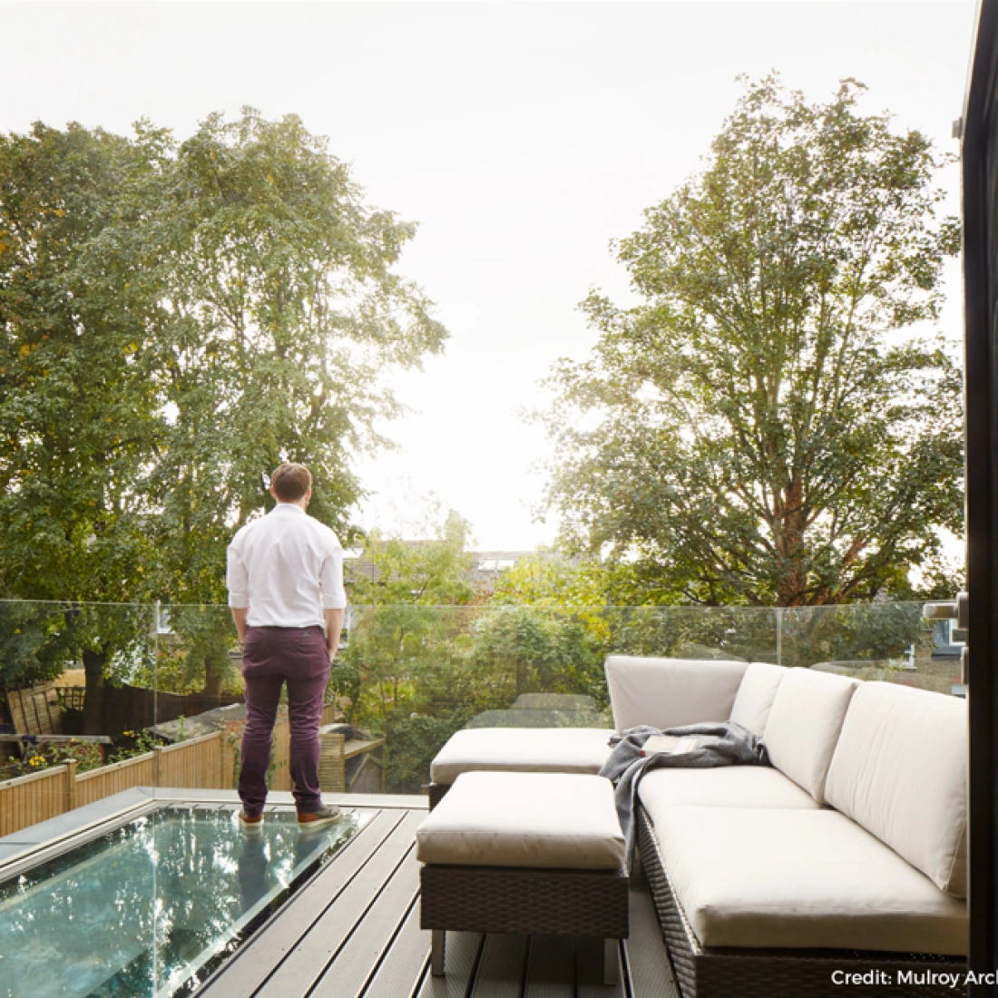 A man stands on a balcony, looking out at trees and a cloudy sky. The balcony features a modern outdoor sofa and a cozy throw blanket.