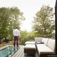 Un homme debout sur un balcon, regardant un paysage verdoyant avec des arbres. Il porte une chemise blanche et un pantalon violet. Un canapé moderne est visible à proximité.