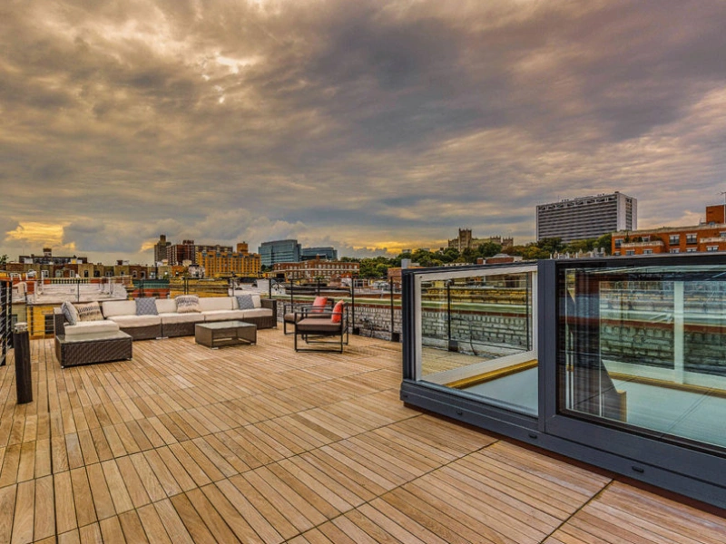 A spacious rooftop terrace featuring wooden flooring, outdoor seating arrangements, and glass railings. The skyline includes buildings and a cloudy sky in the background.