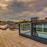 A spacious rooftop terrace with wooden flooring, featuring modern outdoor furniture, including sofas and a dining set. The background showcases a cloudy sky and a city skyline with buildings and a river.