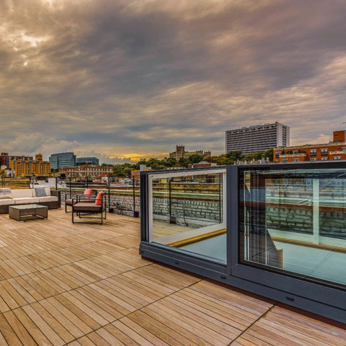 A spacious rooftop terrace with wooden flooring, featuring modern outdoor furniture, including sofas and a dining set. The background showcases a cloudy sky and a city skyline with buildings and a river.