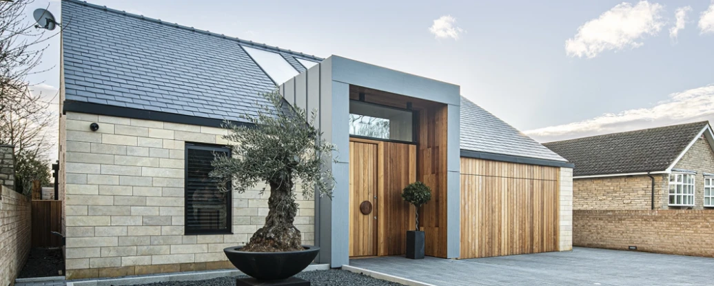 Modern house with a blend of stone and wooden accents, featuring a large front door and a sleek roofline. A decorative olive tree in a black pot is positioned in front, with a paved driveway leading up to the entrance. Mountainous landscape and blue sky in the background.