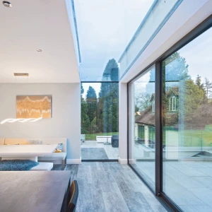 A modern dining area featuring a sleek table and a built-in bench with cushions. Large glass walls provide a view of the outdoor landscape, while a stylish light fixture hangs above the table. Natural light floods the space, enhancing the contemporary design.