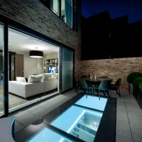 Terraza moderna con piso de vidrio iluminado, mesa y sillas, y una sala de estar visible a través de grandes ventanales. El ambiente es nocturno, con una iluminación suave y un diseño elegante.