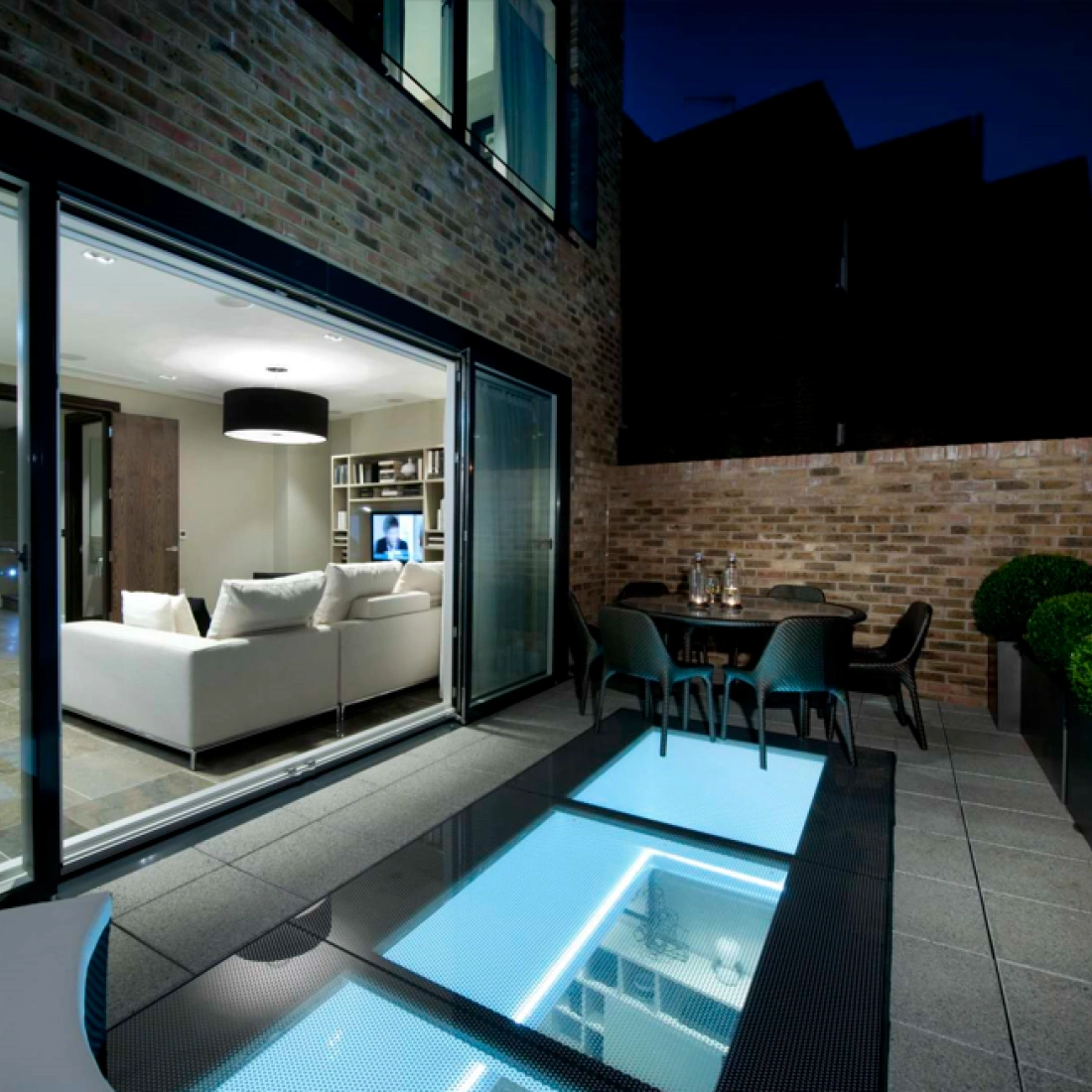 A modern patio area at night features a glass panel floor revealing blue lighting underneath. It opens up to a cozy living room with a light sofa and contemporary decor. Potted plants and a dining table complete the elegant outdoor space.