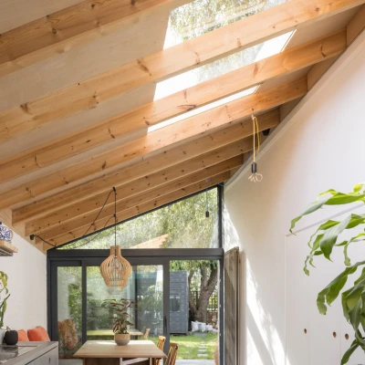 A modern kitchen interior featuring a skylighted, wooden-beamed ceiling. The kitchen has a minimalist design with a metal shelf, potted plants, and a dining table with wooden chairs. Natural light floods the space, illuminating the greenery outside through large glass doors.