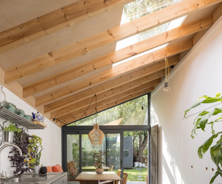 A modern kitchen interior featuring a skylighted, wooden-beamed ceiling. The kitchen has a minimalist design with a metal shelf, potted plants, and a dining table with wooden chairs. Natural light floods the space, illuminating the greenery outside through large glass doors.