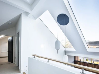 Bright, modern hallway featuring a white exterior, large skylights, and a wooden railing. The minimalist design includes angular architectural elements and a round pendant light.