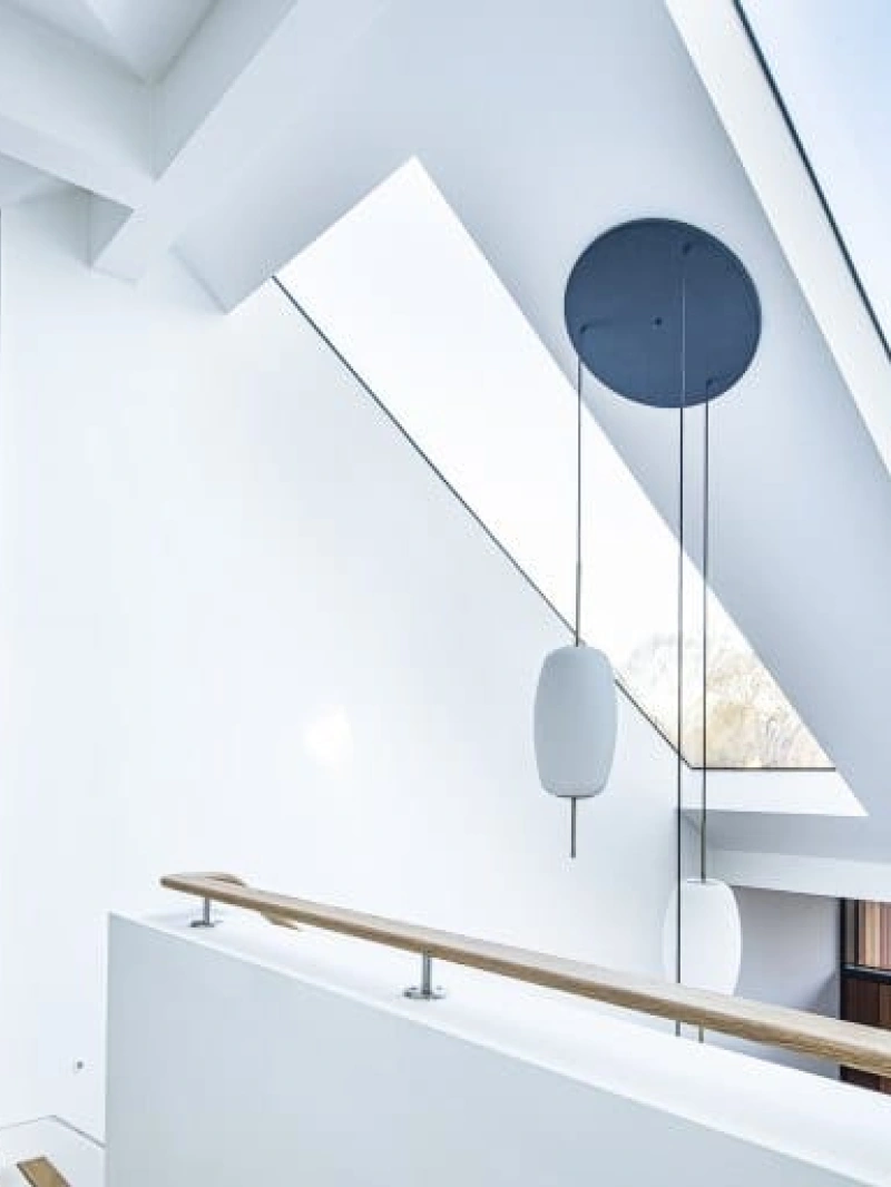 A modern, well-lit hallway with a sloped ceiling and large skylights. A sleek, minimalistic hanging light fixture is suspended from the ceiling. The walls are painted white, and the railing is made of wood.
