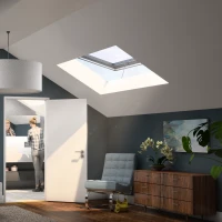A modern attic bedroom featuring a large bed with crumpled white linens, a sleek bedside lamp, and a wooden dresser. Two skylights illuminate the space, and a person is visible in the bathroom through an open door. Abstract art hangs on the walls, and a potted plant adds a touch of greenery.