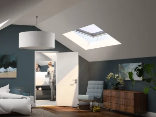 Bright and modern attic bedroom featuring a bed with white bedding, a stylish bedside lamp, and artwork on the walls. A woman is seen in the adjoining bathroom, while large skylights let in natural light. A wooden dresser and a contemporary chair complete the cozy atmosphere.