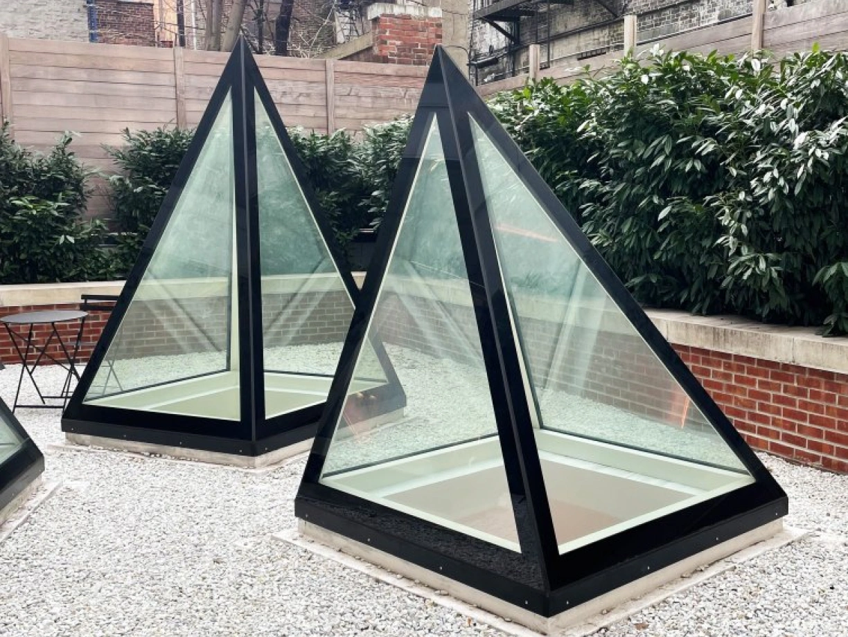 Two modern glass and black-frame pyramidal structures are placed on a gravel surface in a courtyard, surrounded by low greenery and brick walls. An apartment building with fire escapes is visible in the background.