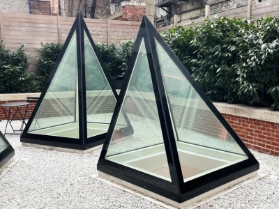 Two modern glass pyramids with black frames sit in a gravel garden area, surrounded by greenery and brick walls. In the background, brick buildings with fire escapes are visible.