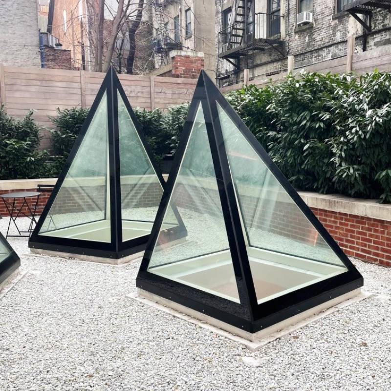 Two modern glass pyramids with black frames sit in a gravel garden area, surrounded by greenery and brick walls. In the background, brick buildings with fire escapes are visible.