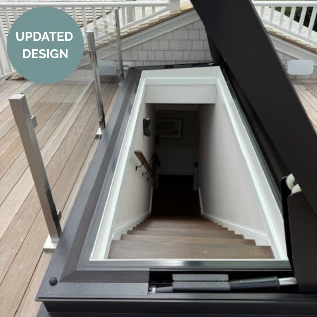 A stairwell leading down from a rooftop, featuring wooden steps and a light-colored wall. The opening is framed with modern materials, and a glass railing is visible at the side. A circular label in the top left corner reads "UPDATED DESIGN."