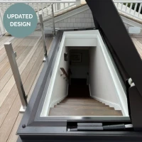 A stairwell leading down from a rooftop, featuring wooden steps and a light-colored wall. The opening is framed with modern materials, and a glass railing is visible at the side. A circular label in the top left corner reads "UPDATED DESIGN."