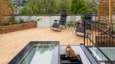 A rooftop garden featuring wooden decking, two black lounge chairs, a small table, and a cat sitting near a set of stairs that lead to a lower level. Lush greenery surrounds the area with buildings in the background.