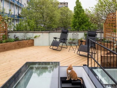 Terraza con piso de madera, dos sillas reclinables negras y una mesa. Hay un gato anaranjado sentado cerca de la entrada de una escalera. En el fondo, se ven árboles y edificios.