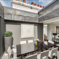 A modern dining area featuring a sleek dark table surrounded by white chairs. A large abstract painting hangs on the wall, with greenery in a tall planter nearby. Large windows allow natural light to fill the space, which has a contemporary design aesthetic.