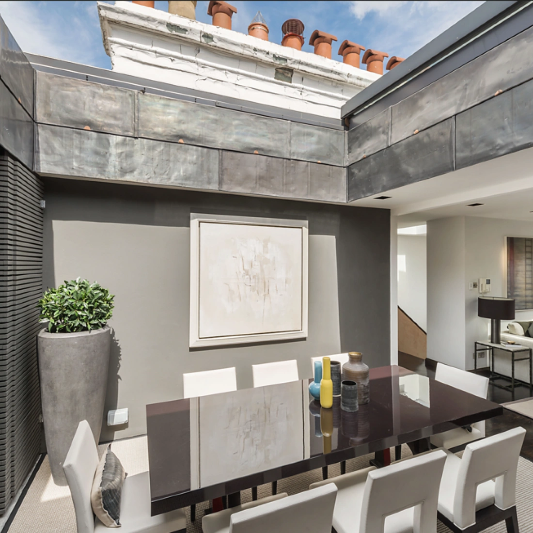 A modern dining area featuring a sleek dark table surrounded by white chairs. A large abstract painting hangs on the wall, with greenery in a tall planter nearby. Large windows allow natural light to fill the space, which has a contemporary design aesthetic.