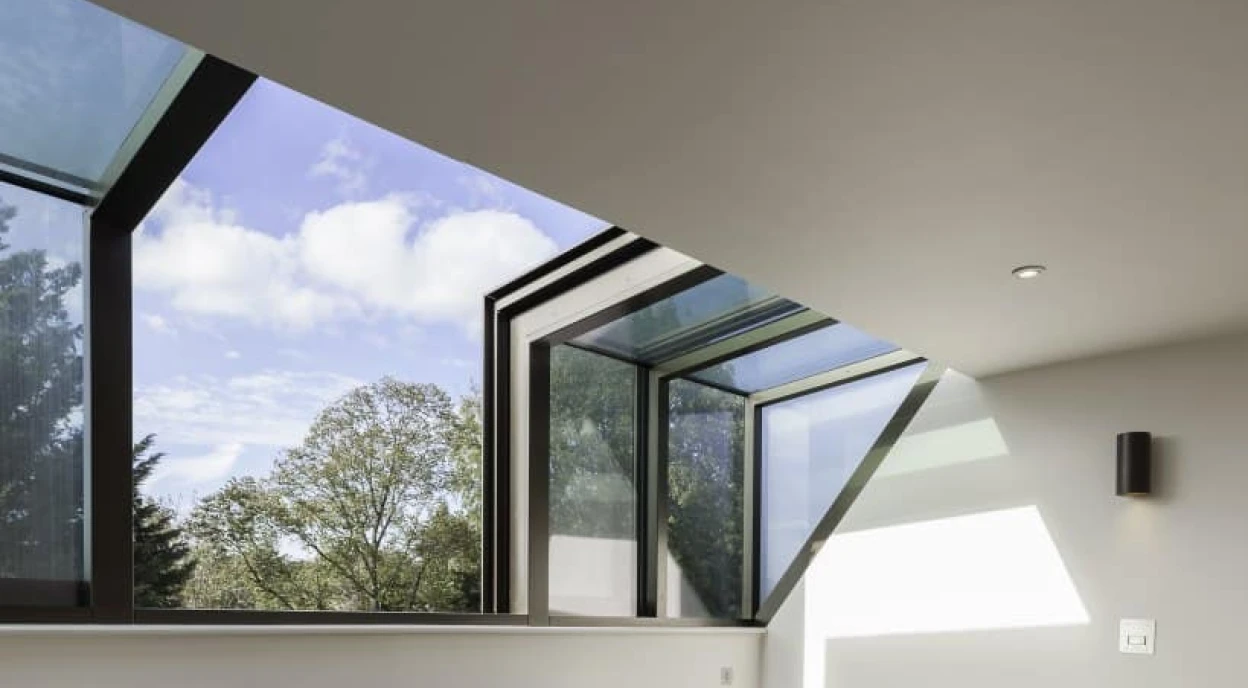 Deux fauteuils bleus sont placés dans une pièce lumineuse avec de grandes fenêtres triangulaires. Le sol est en bois clair et des rayons de lumière traversent la pièce, créant des ombres intéressantes. Des arbres sont visibles à l’extérieur.