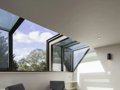 Two blue upholstered armchairs sit on a wooden floor in a bright room with large windows. The angular window design allows natural light to fill the space, and trees are visible outside. A small shelf is located beside the chairs.