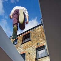 Una persona con pantalones oscuros y zapatos claros parece estar caminando sobre un tragaluz, con una vista del cielo azul y nubes. La pared de ladrillo expuesta de una casa se ve detrás.