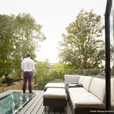 A person stands on a balcony overlooking a lush green landscape. The balcony features a glass railing and a cozy seating area with light-colored cushions. The sky is bright, indicating daylight.