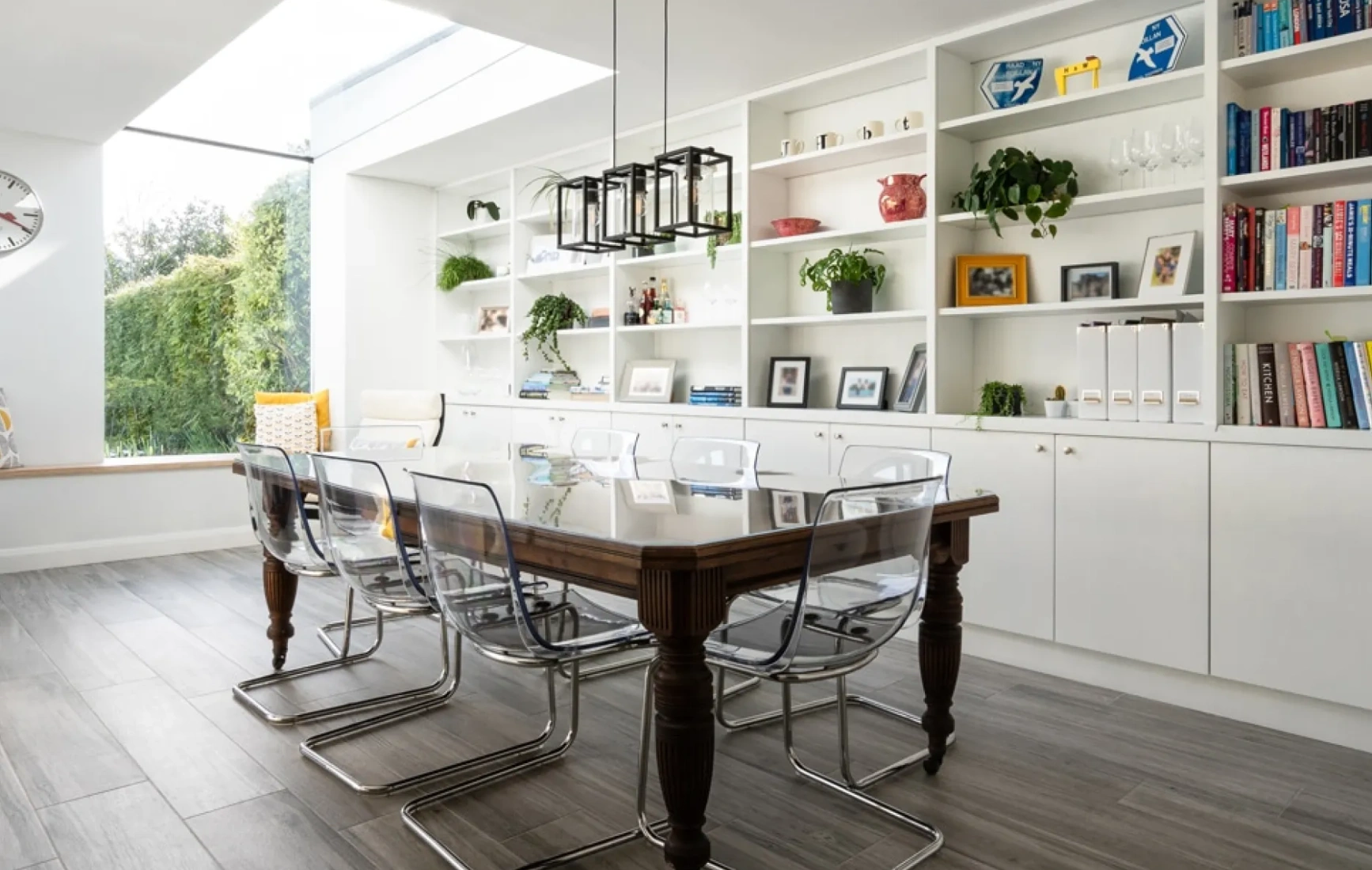 A modern dining area with a large wooden table and transparent chairs. White shelves filled with books, plants, and decorative items line the walls. A skylight allows natural light to flood in, creating a bright and airy atmosphere.