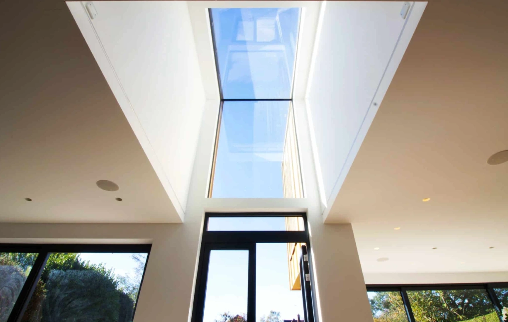 Bright modern interior featuring a large skylight that brings natural light into the space. The view through the skylight shows a clear blue sky. Large glass doors open to a green outdoor area.