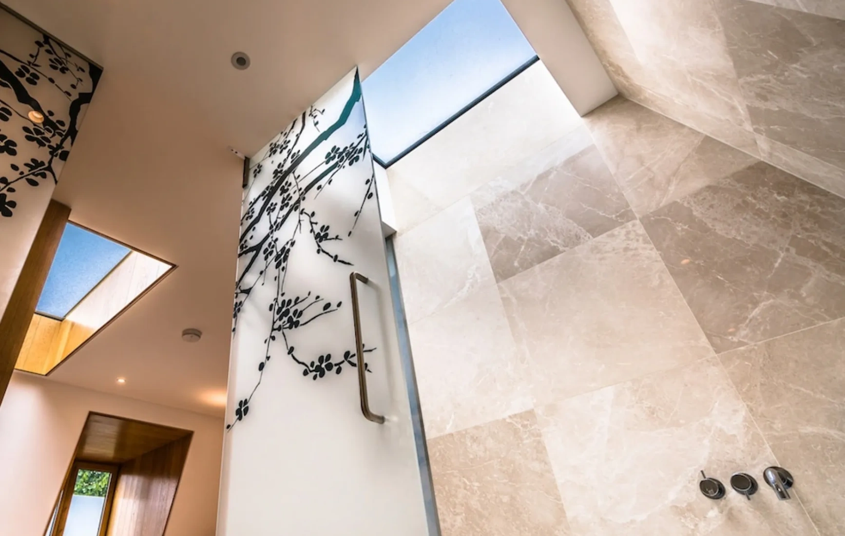 A modern bathroom featuring large, marble tiles and a frosted glass door with a floral design. Natural light streams in through skylights, illuminating the space.