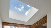 A modern skylight set in a ceiling, revealing a bright blue sky with a few fluffy clouds. Natural wood framing complements the minimalist design.