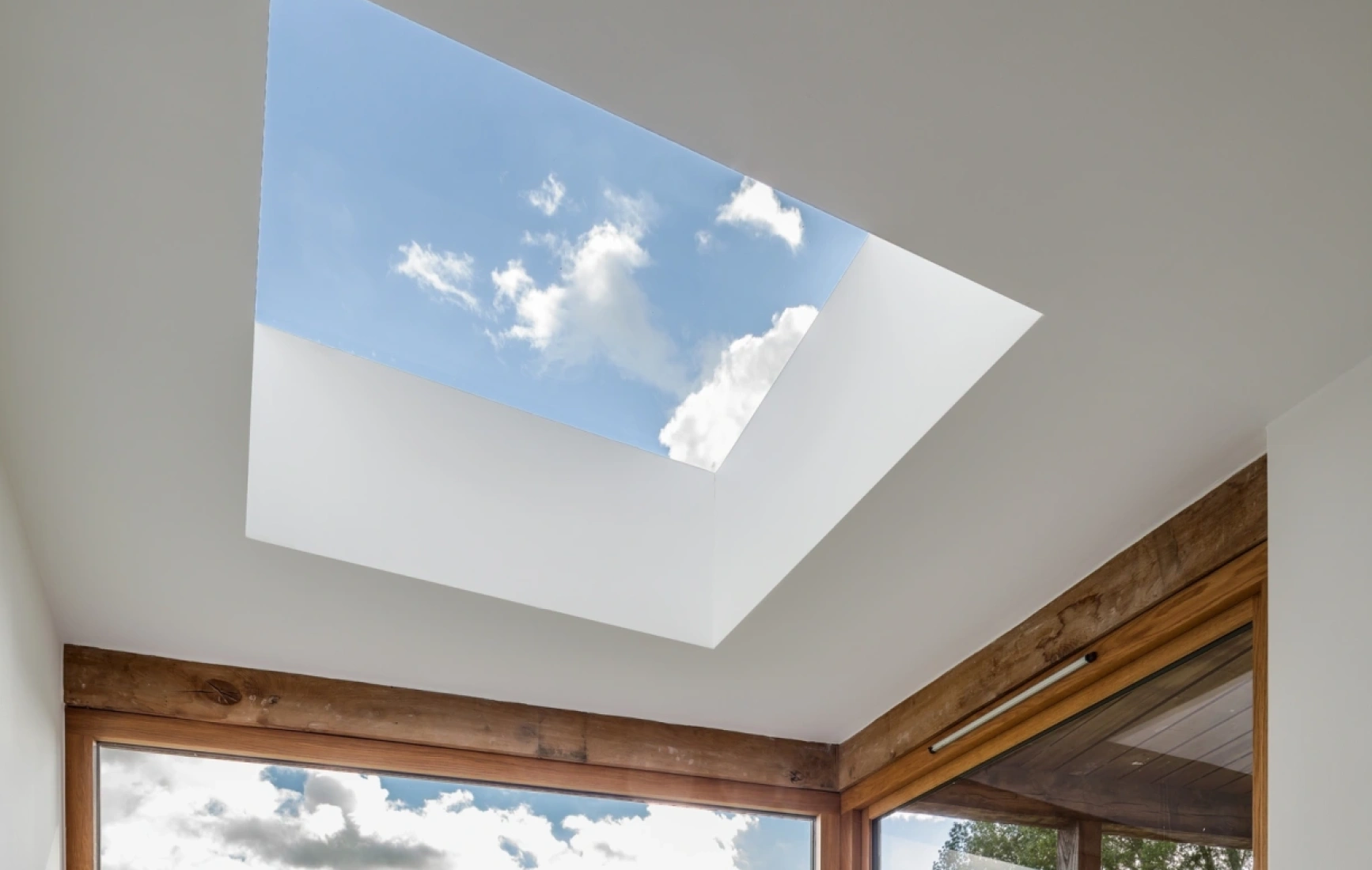 A modern skylight set in a ceiling, revealing a bright blue sky with a few fluffy clouds. Natural wood framing complements the minimalist design.