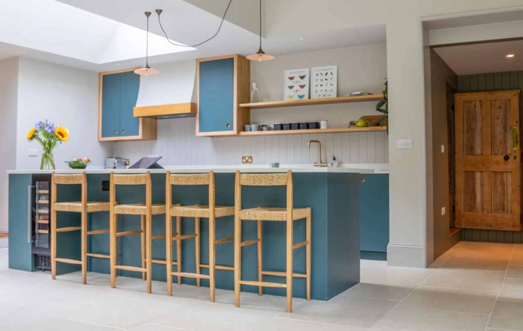 A modern kitchen featuring a large island with four wooden bar stools. The island has a sink and is surrounded by light blue cabinets. Sunflowers are displayed in a vase on the countertop, with a wooden door visible in the background. Natural light floods the space through a skylight.