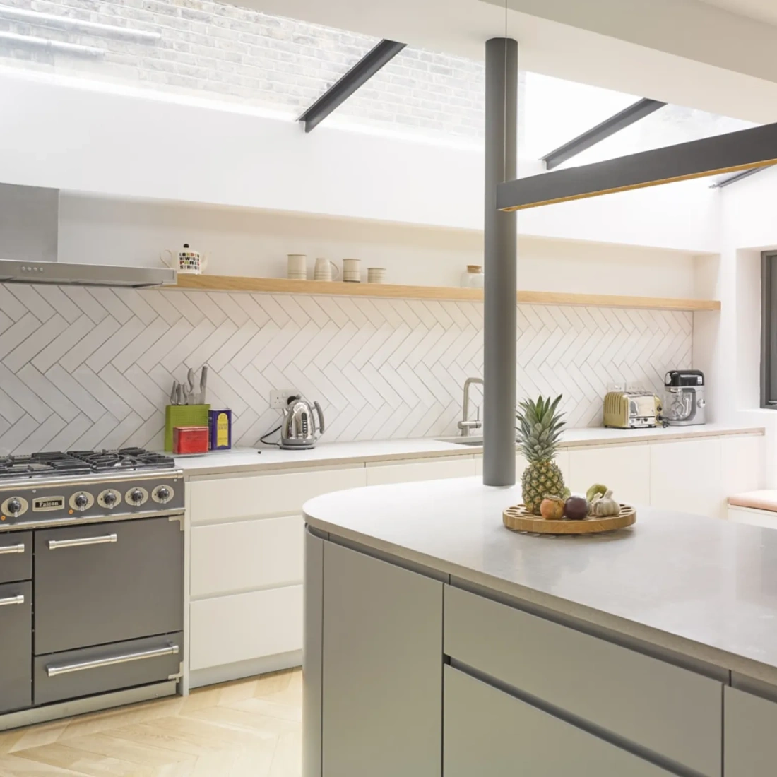 Cucina moderna con paredes bianche, armarios minimalisti e pavimento in legno. Un forno a gas grigio e un piano di lavoro in marmo. Un piatto con un'ananas e frutta su un piano. Finestroni che lasciano entrare abbondante luce naturale.
