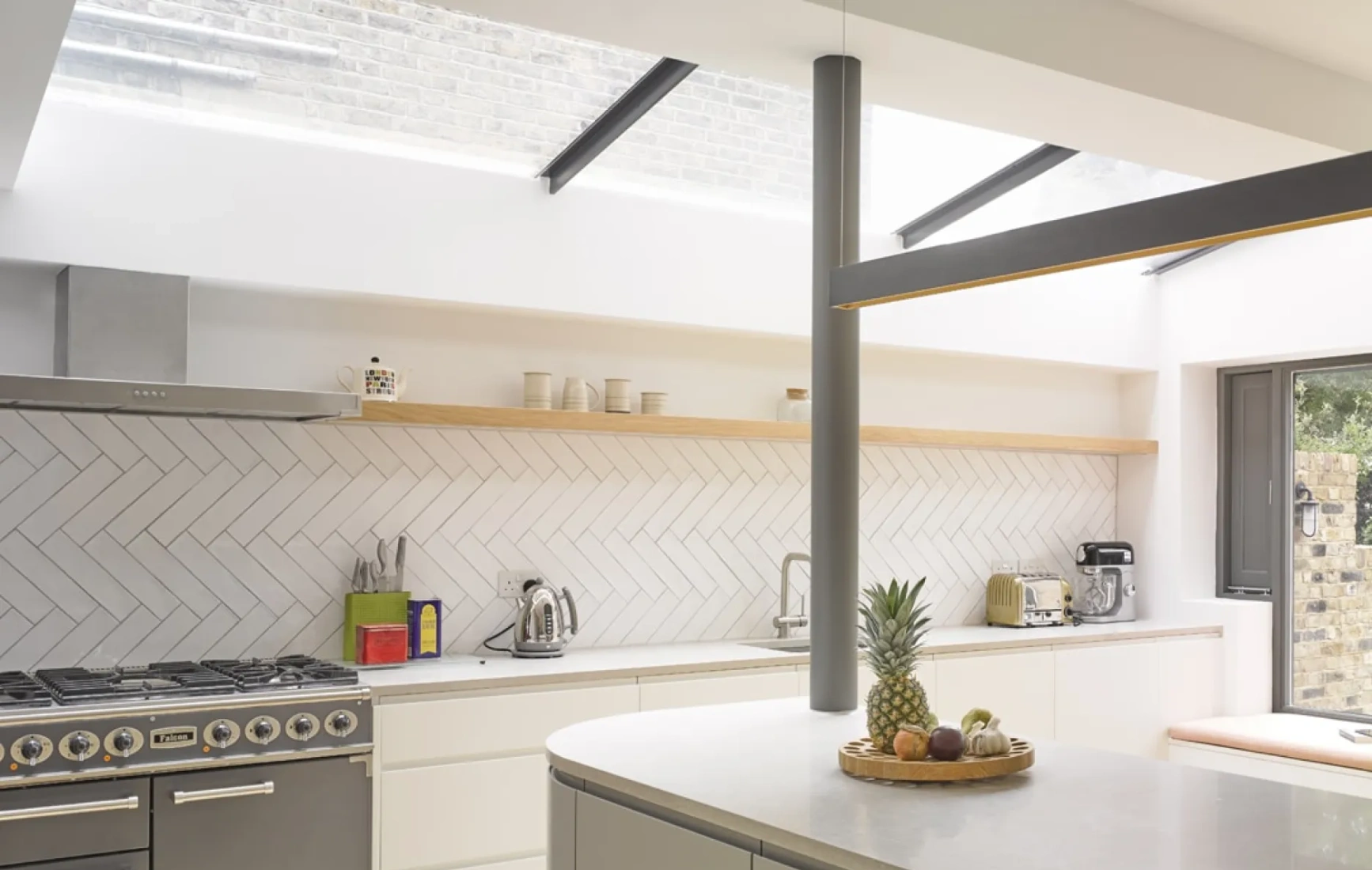 A modern kitchen featuring a gray range cooker, herringbone tiled walls, and a light-filled space with open shelving. A countertop displays a wooden tray with fruits, and large windows provide ample natural light.