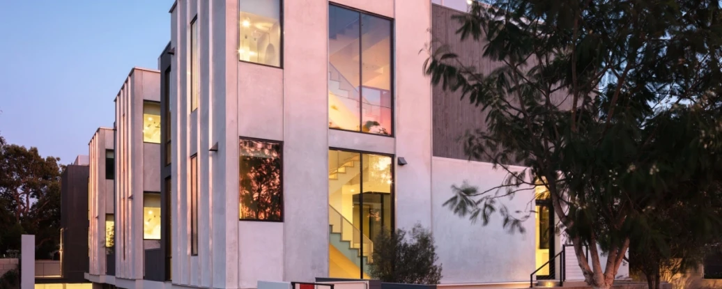Modern multi-story building with large glass windows and a sleek exterior, illuminated in the evening. The architecture features a mix of light and dark stucco, with a landscaped entrance and outdoor lighting.
