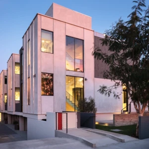 Modern multi-story building with large glass windows and a sleek exterior, illuminated in the evening. The architecture features a mix of light and dark stucco, with a landscaped entrance and outdoor lighting.